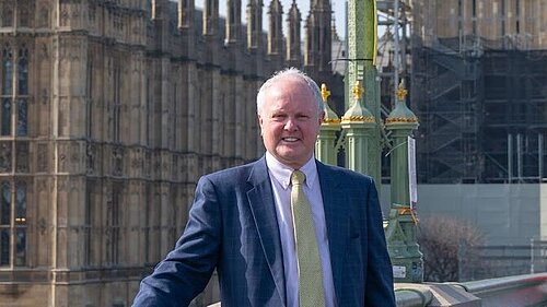 Clive Jones MP in a suit with a yellow tie. He is stood on Westminster Bridge with the palace of Westminster in the background. He is smiling at the camera.